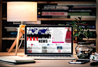 Laptop displaying fake news graphic on desk with lamp and plant