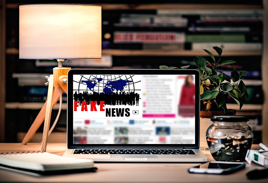 Laptop displaying fake news graphic on desk with lamp and plant
