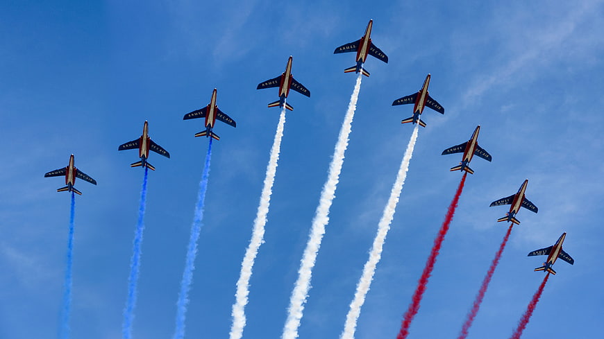 French Air Force Patrouille de France jets flying in formation with red, white, and blue smoke trails against a blue sky.