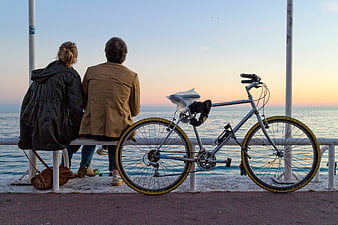 Couple sitting on bench by sea with bicycle at sunset
