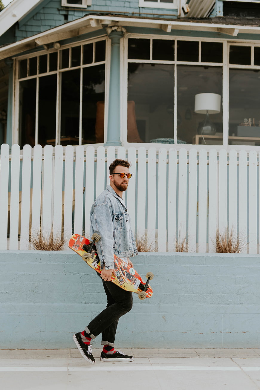 Man with skateboard walking past blue house and white fence