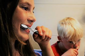 Girl brushing teeth with boy in background bathroom