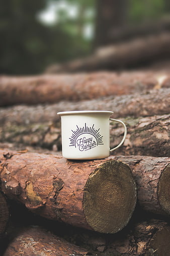 White enamel mug with Happy Camper text on forest logs