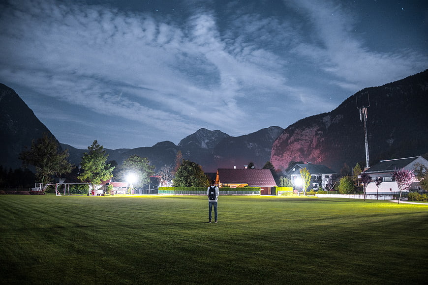 Lone Man Standing on Illuminated Grass Field at Night with Mountains