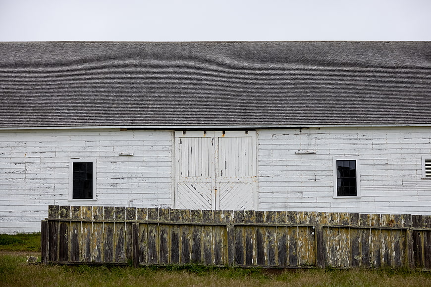 Weathered white wooden barn with rustic fence and gray roof