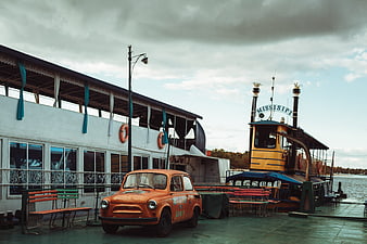 Vintage orange car parked on a dock near a riverboat
