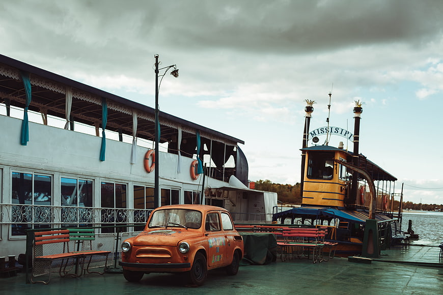 Vintage orange car parked on a dock near a riverboat