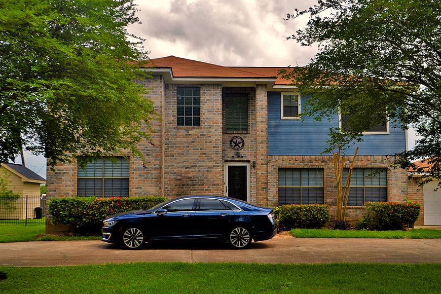 Blue Lincoln MKZ sedan parked in front of a two-story brick and blue siding suburban house