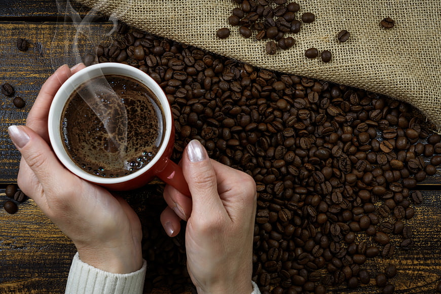 Hands holding steaming coffee cup over roasted beans