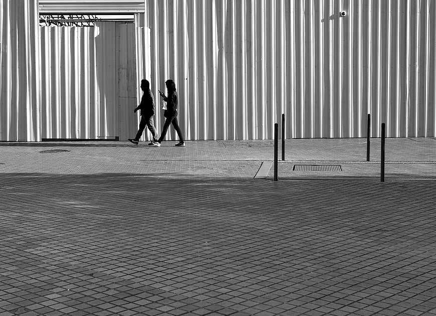 Two people walking past corrugated metal wall in black and white