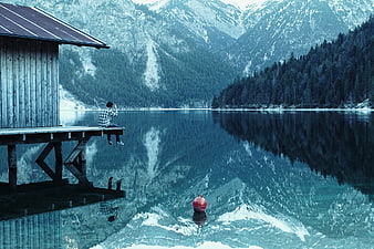 Man sitting on wooden pier by alpine lake with mountain reflections