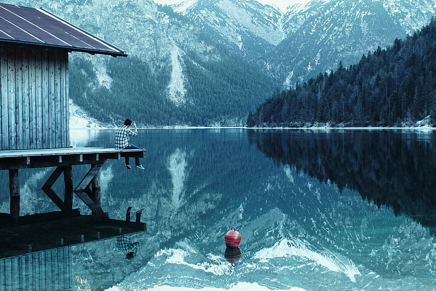Man sitting on wooden pier by alpine lake with mountain reflections
