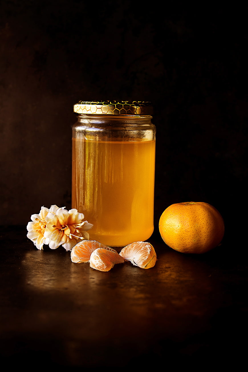 Glass Jar of Honey with Tangerines and Flowers on Dark Background