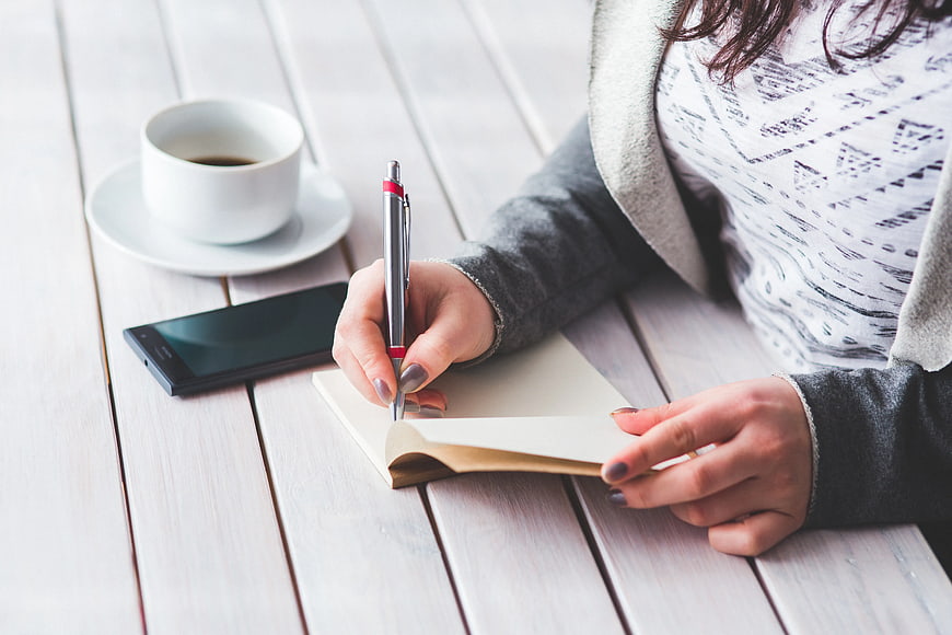 Woman writing in notebook with coffee and smartphone