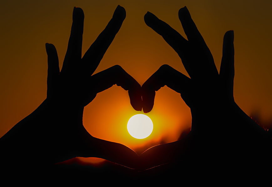 Silhouette of hands forming a heart shape around a bright sunset sun