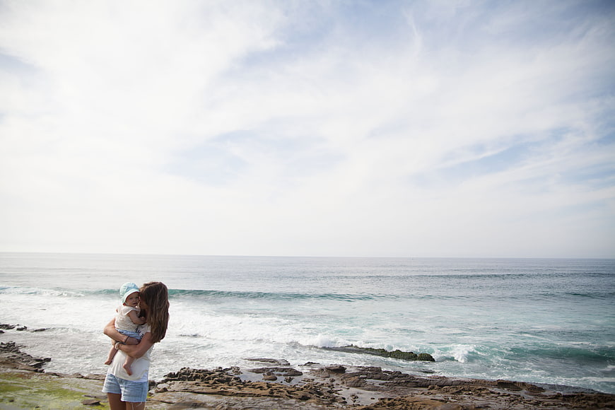 Mother holding baby on rocky beach looking at ocean