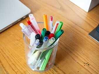Colorful pens and markers in a glass cup on a wooden desk