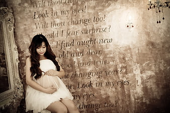 Pregnant woman in white dress sitting against textured wall with text