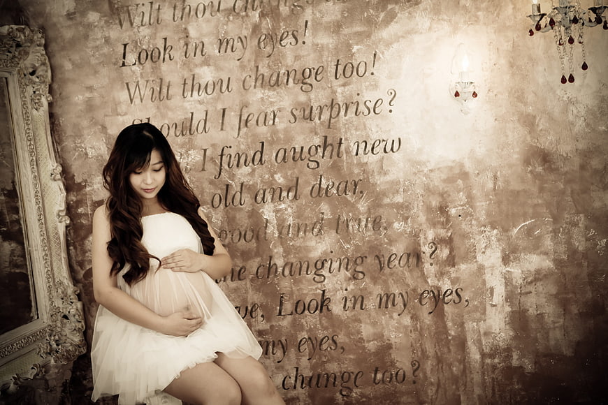 Pregnant woman in white dress sitting against textured wall with text