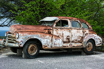 Rusty vintage car wreck on pavement with green trees