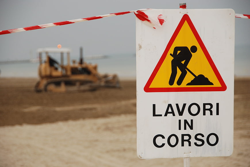 Italian Road Works Sign With Blurred Construction Vehicle On Beach