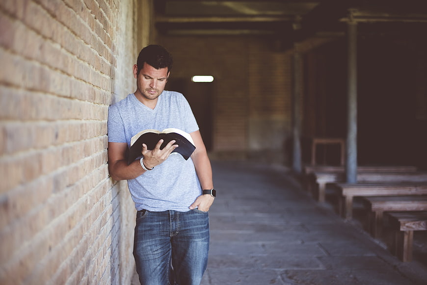 Man reading book leaning against brick wall in hallway