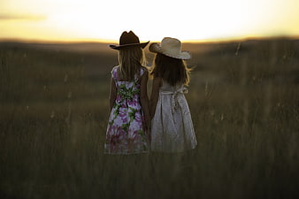 Two young girls in cowboy hats holding hands in a field at sunset