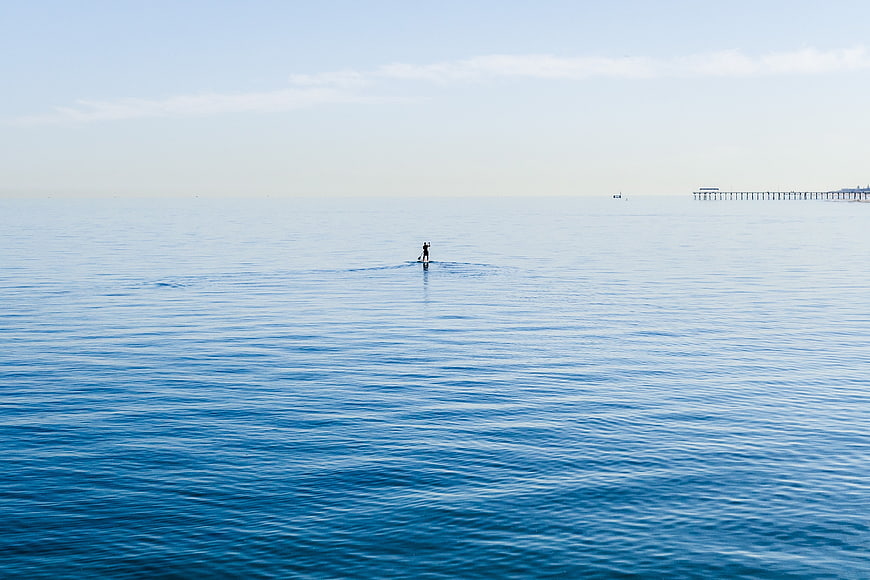 Lone paddleboarder on calm blue ocean with distant pier