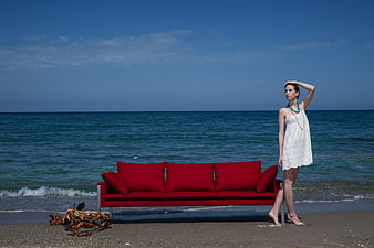 Woman in white dress standing next to red sofa on beach