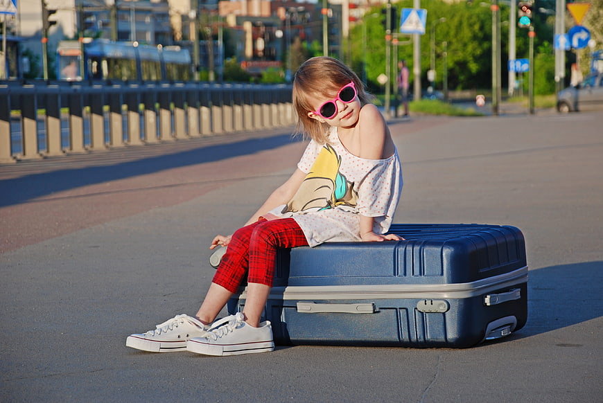 Little girl in pink sunglasses sitting on blue suitcase in city street