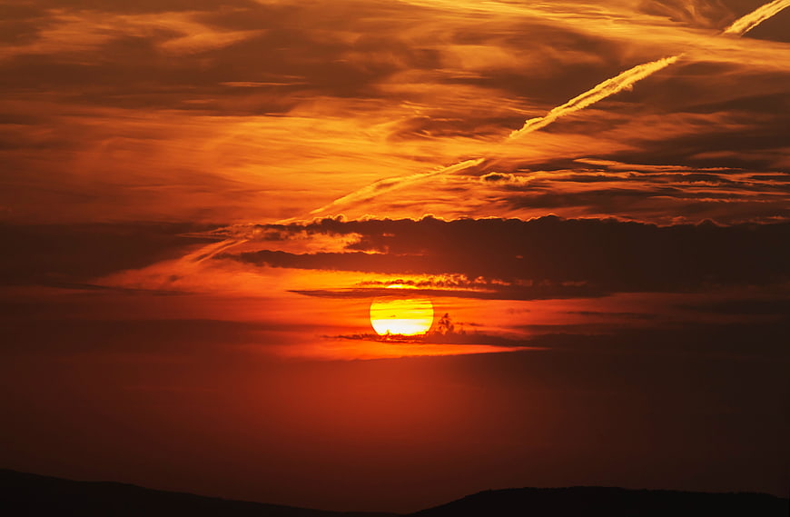 Vibrant orange sunset with clouds and contrails over dark hills
