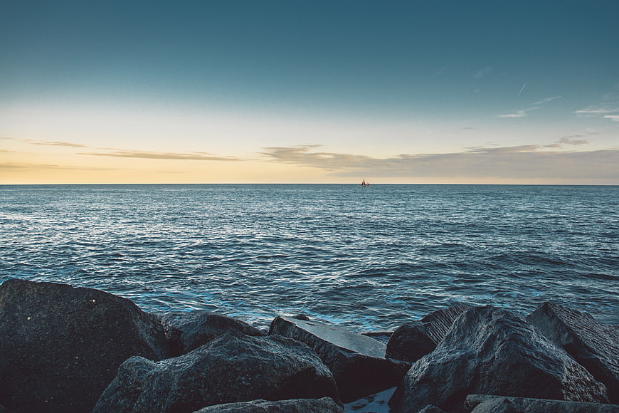 Serene Ocean Sunset with Distant Sailboat and Foreground Rocks