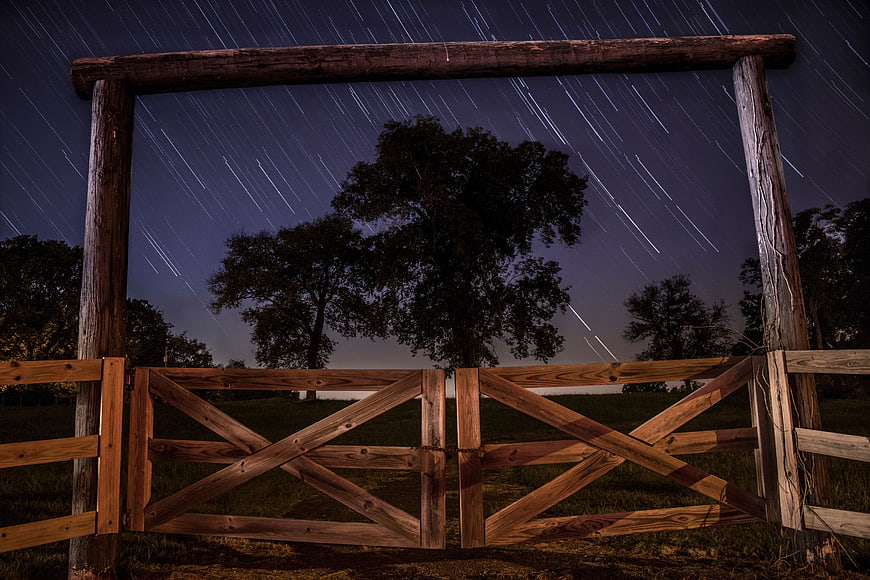 Star trails over rustic wooden gate and trees at night