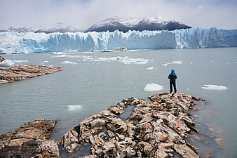 Hiker standing on rocky shore facing massive glacier and mountains