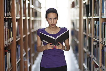 Woman reading book in library aisle looking at camera