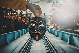 Hand holding black Guy Fawkes mask over railway tracks at sunset