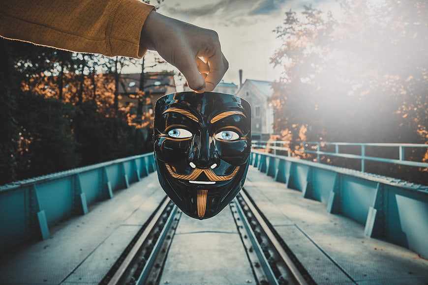Hand holding black Guy Fawkes mask over railway tracks at sunset