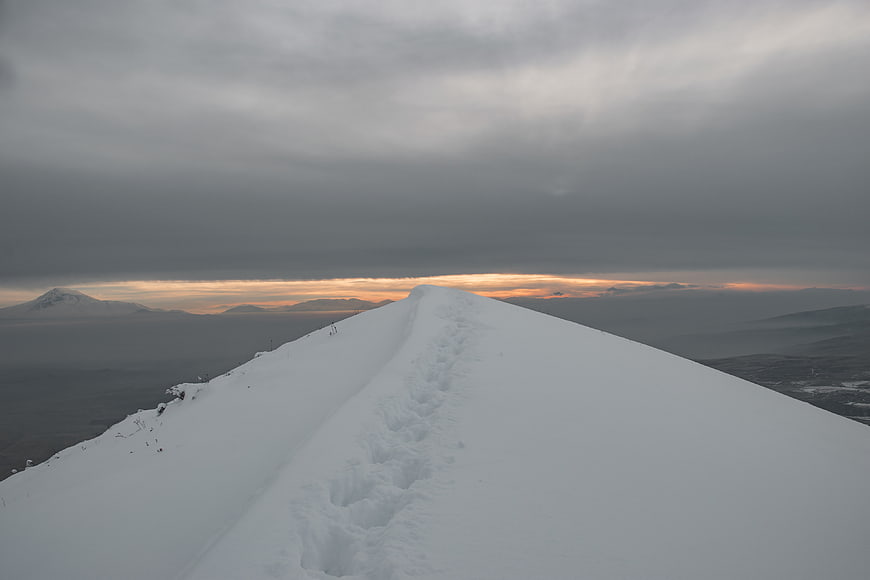 Snowy mountain ridge with footprints leading to sunset horizon