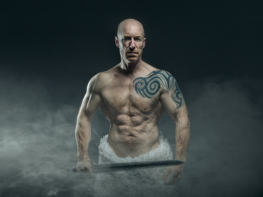 Shirtless muscular man with tattoo holding sword in dramatic smoke