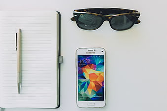 Flat lay of notebook, pen, smartphone, and sunglasses on white desk