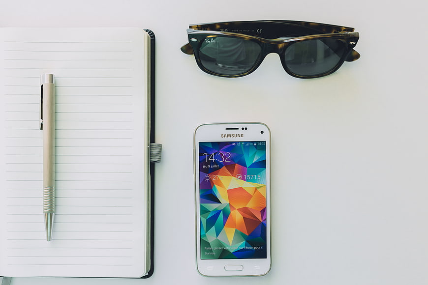 Flat lay of notebook, pen, smartphone, and sunglasses on white desk