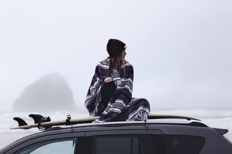 Woman wrapped in blanket sitting on car roof with surfboard in foggy ocean