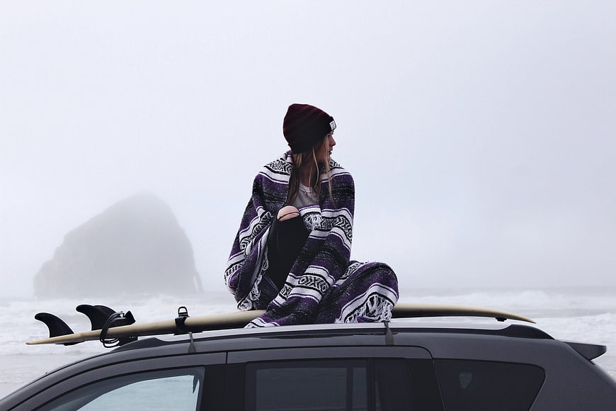 Woman wrapped in blanket sitting on car roof with surfboard in foggy ocean