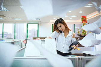 Stressed Asian businesswoman on phone in busy office with flying papers