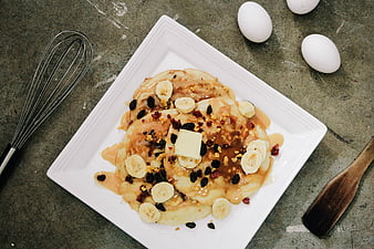 Overhead view of pancakes with bananas, raisins, and butter on a white plate