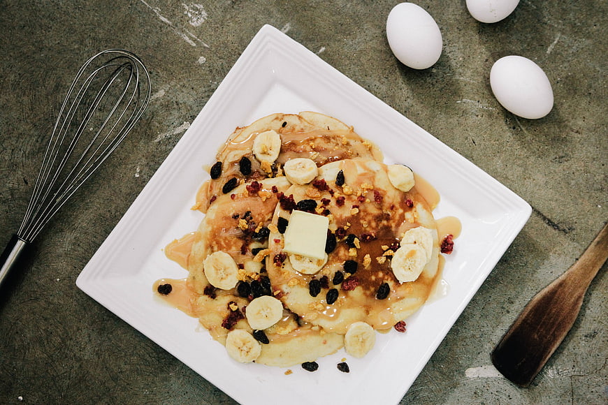Overhead view of pancakes with bananas, raisins, and butter on a white plate