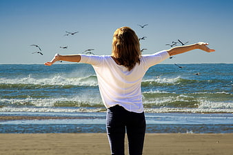 Woman with open arms on sunny beach facing ocean waves and flying seagulls