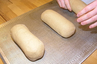 Hands shaping brown bread dough on perforated baking sheet