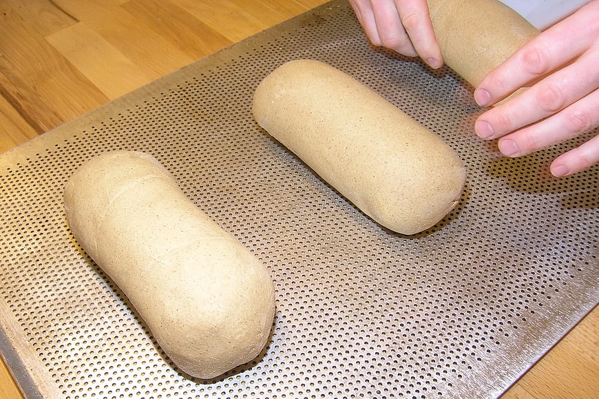 Hands shaping brown bread dough on perforated baking sheet