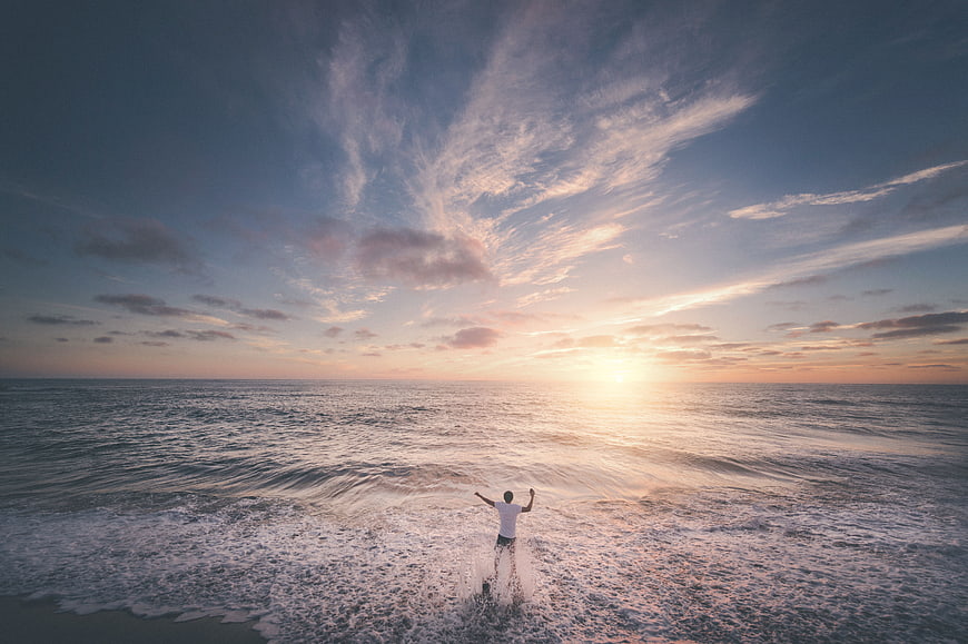 Man with arms raised in ocean waves at sunset